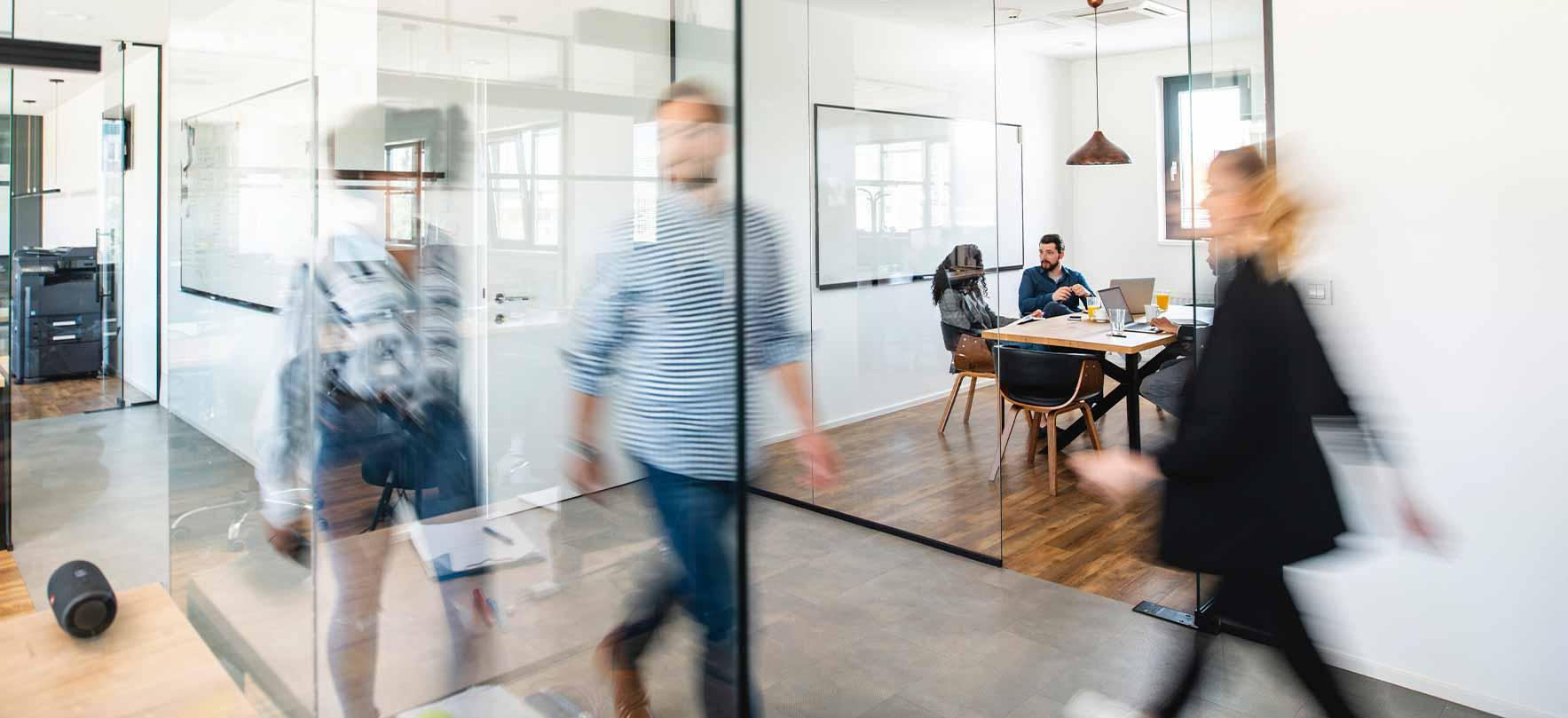 People walking through a modern office while a team meets in a glass-walled meeting room