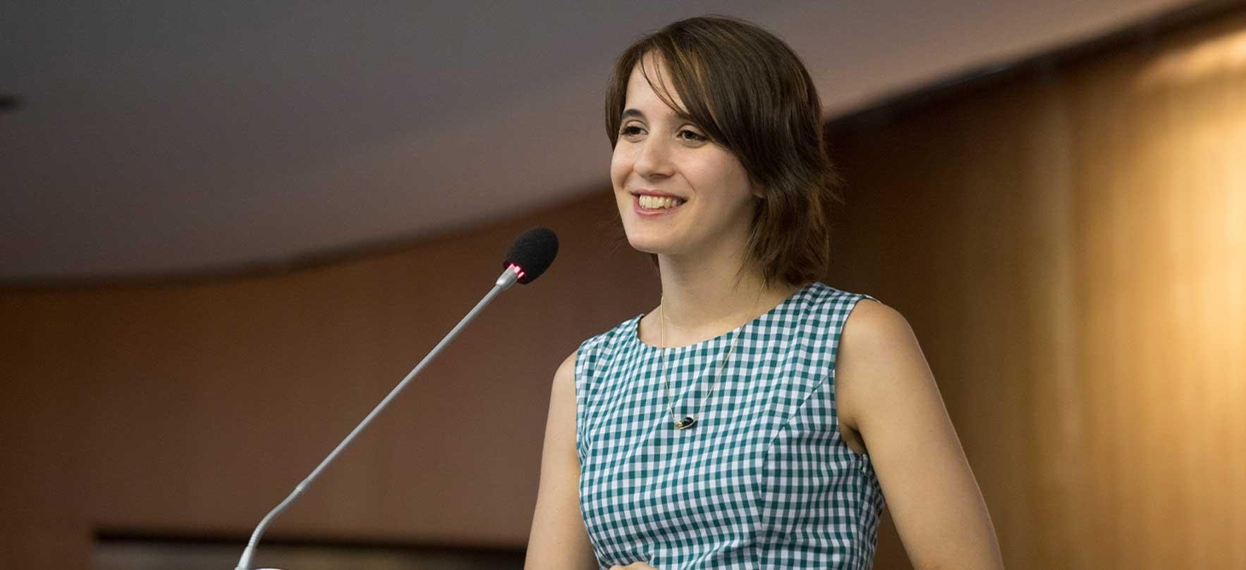 Smiling young woman speaking at a podium with a microphone during a formal event or conference.