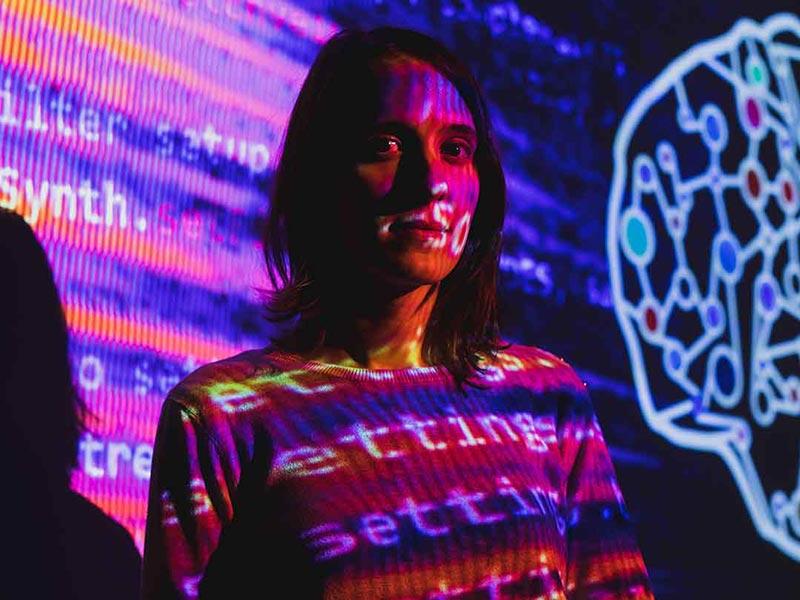 Woman standing in front of projected code and a digital brain illustration, with colorful light patterns on her face